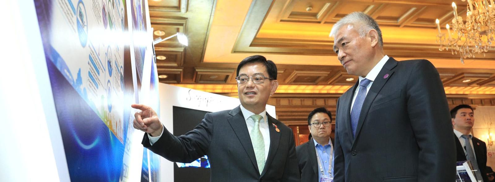 Two men in suits view charts on a poster; one points at the graphics.